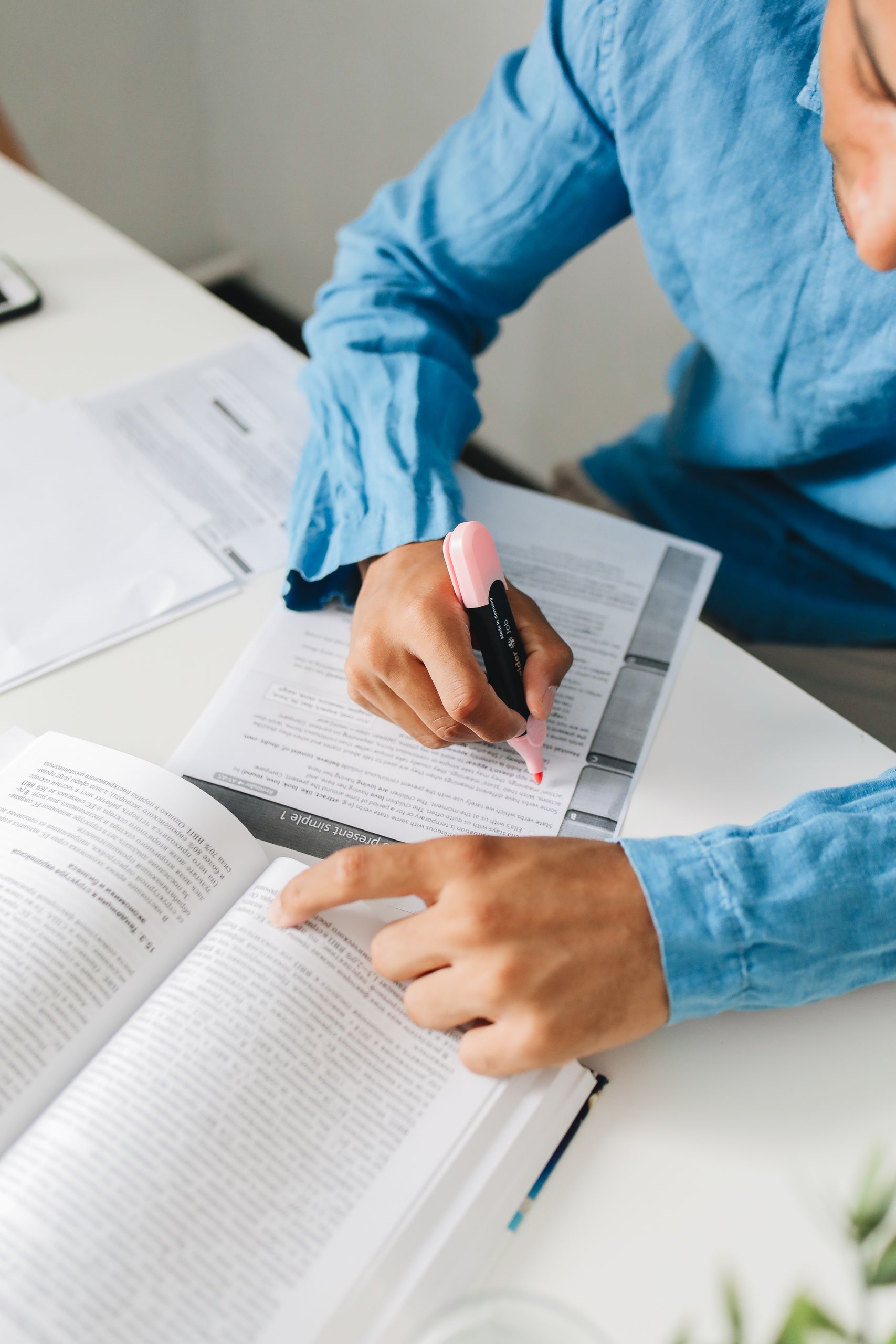 A person in blue shirt highlighting text in a book with a pen while studying indoors.